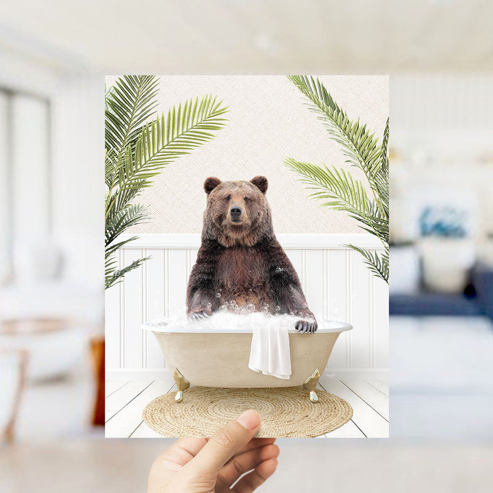A hand is holding up a framed image of a brown bear sitting in a bathtub filled with bubbles, surrounded by green palm leaves.