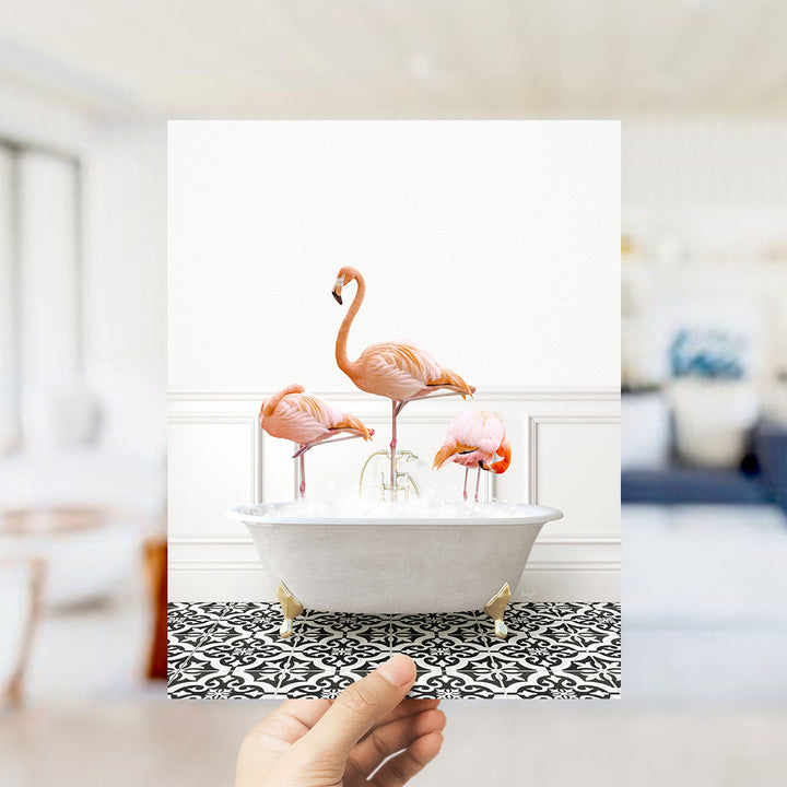 A hand holds up a framed image of three flamingos standing in a white bathtub filled with water.