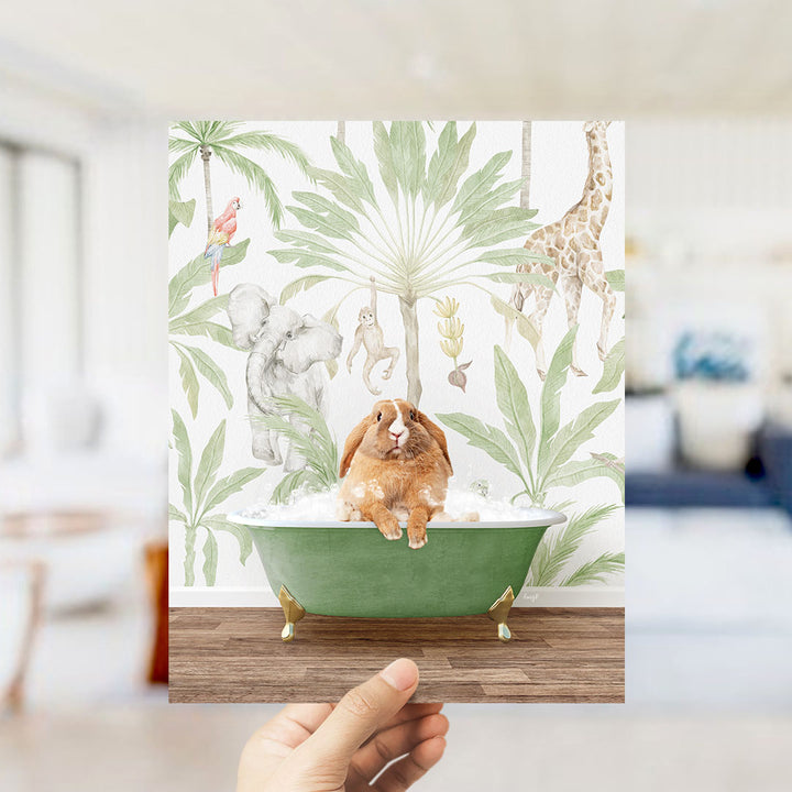 A hand holds up a framed print featuring a whimsical scene of a rabbit sitting in a green bathtub surrounded by various animals, including a giraffe, elephant, and parrot, all set against a tropical wallpaper backdrop.