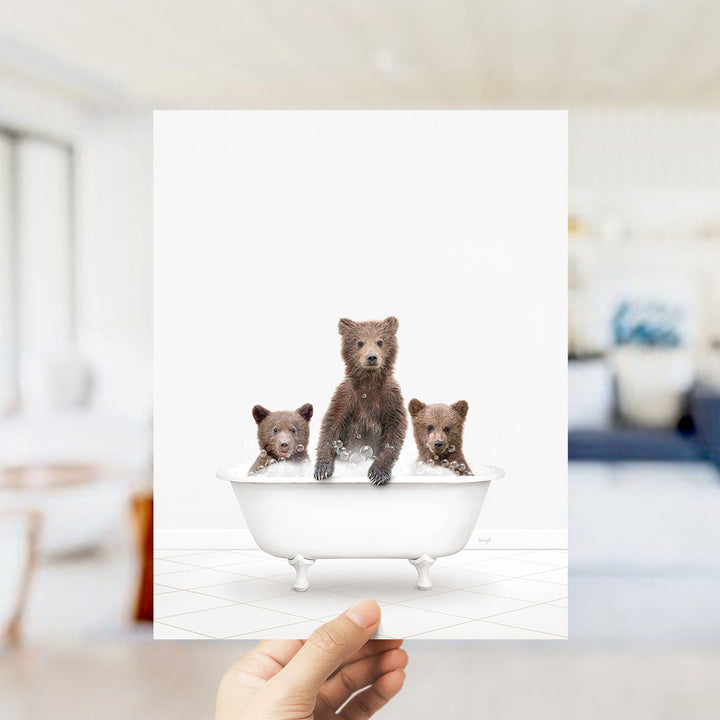A hand holds up a framed image depicting three brown bears sitting in a white bathtub.