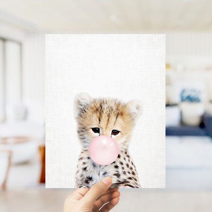 A hand is holding up a framed print of a cheetah blowing a bubble with a pink bubble gum bubble.