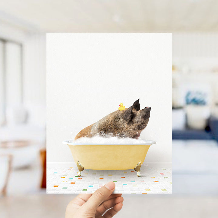 A hand holds up a framed image of a pig sitting in a yellow bathtub, surrounded by colorful tiles.