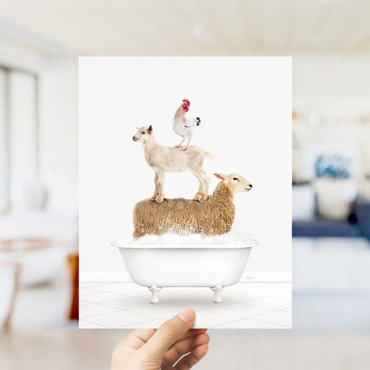 A hand holds up a framed image depicting a goat standing on a sheep's back in a bathtub, with a rooster perched on top of the sheep.