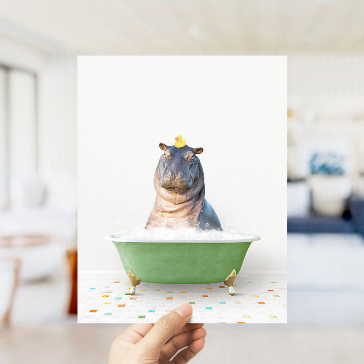 A hand holds up a framed image of a hippopotamus sitting in a green bathtub, with a yellow rubber duck perched on its head.
