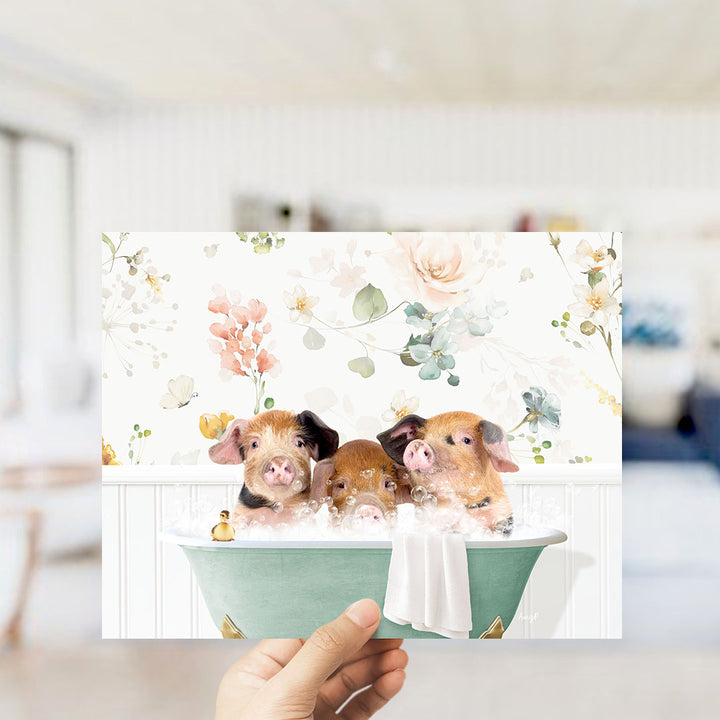 A hand is holding up a framed image of three adorable pigs sitting in a bathtub filled with bubbles, surrounded by a floral pattern.