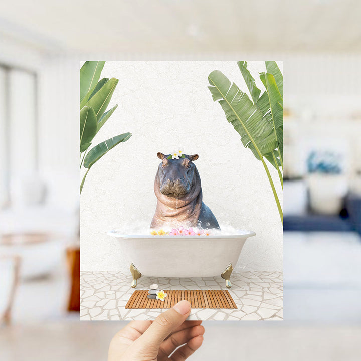 A hand holds up a framed image depicting a hippopotamus sitting in a bathtub filled with bubbles, surrounded by tropical plants.