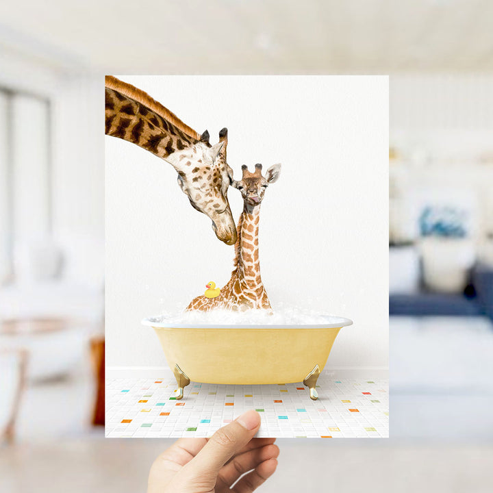 A hand holds up a framed image depicting two giraffes in a yellow bathtub, with one giraffe gently nuzzling the other.