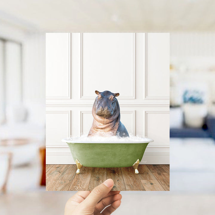 A hand holds up a photograph of a hippopotamus sitting in a green bathtub filled with bubbles, creating a whimsical and playful scene.