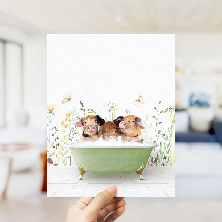 A hand holds up a framed image depicting three adorable pigs sitting in a green bathtub, surrounded by flowers and butterflies.