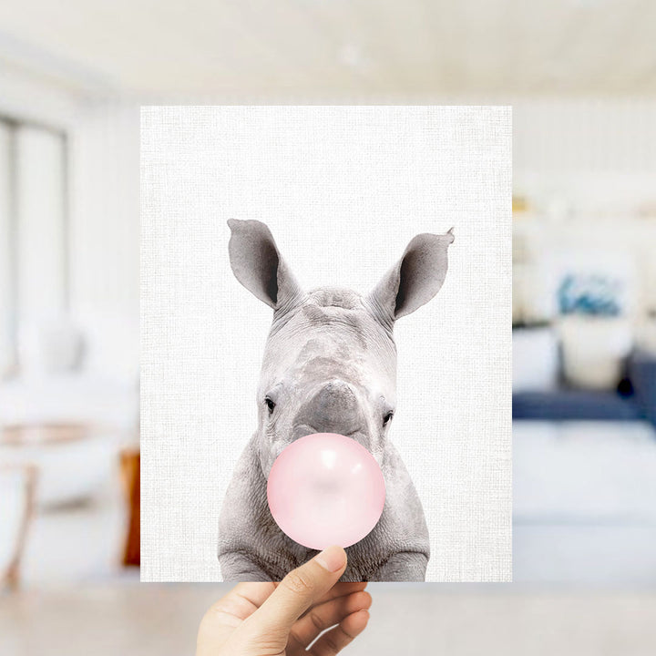 A hand is holding up a framed image of a rhinoceros blowing a bubble with a pink bubble gum bubble.