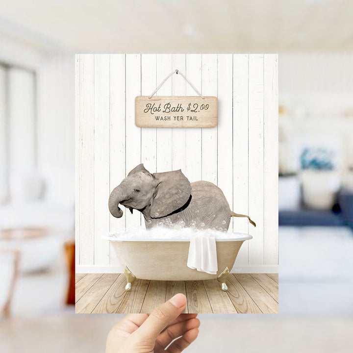 A hand holding a framed image of a baby elephant sitting in a bathtub, with a sign above it that says "Hot Bath $2.00 WASH YOUR TAIL".
