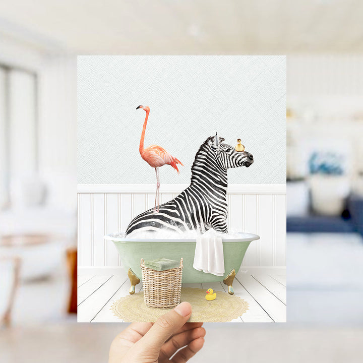 A hand holds up a framed image depicting a zebra sitting in a bathtub, with a pink flamingo standing on its back and a yellow rubber duck floating nearby.