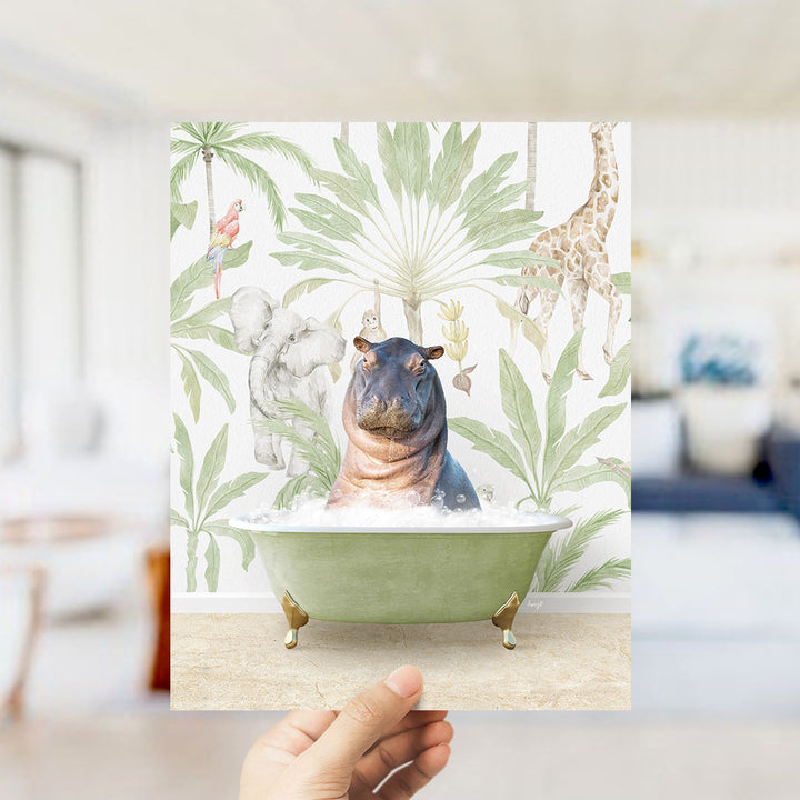 A hand holds up a framed image depicting a hippopotamus sitting in a bathtub surrounded by tropical foliage, including a giraffe and palm trees.