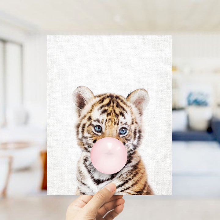 A hand is holding up a framed photograph of a young tiger cub blowing a bubble with a pink bubble gum bubble.
