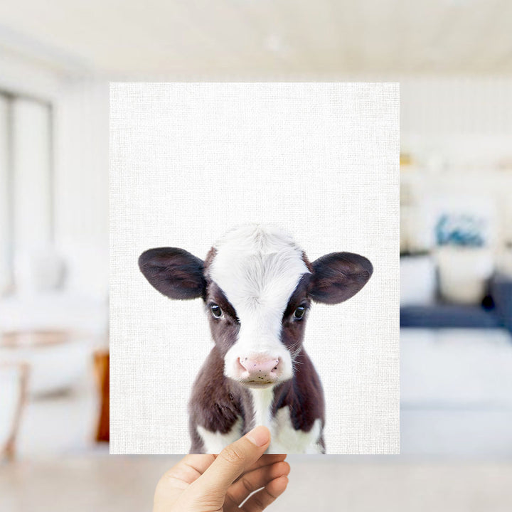 A hand is holding up a white canvas with a close-up of a black and white cow's face.