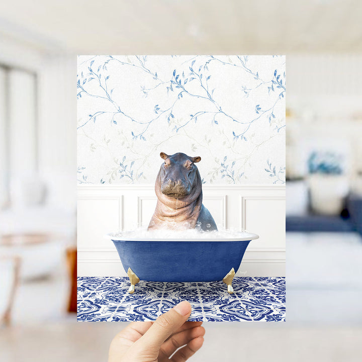 A hand holds up a framed image of a hippopotamus sitting in a blue bathtub, surrounded by a blue and white floral patterned wall.