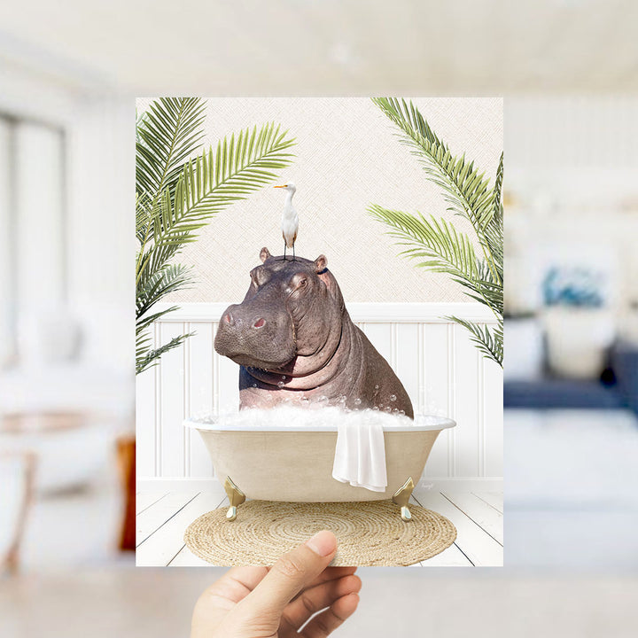 A hand holds up a framed image depicting a hippopotamus sitting in a bathtub, surrounded by palm leaves and a bird.