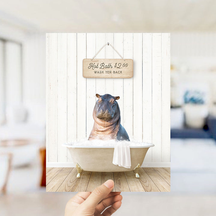A hand holding a framed image of a hippopotamus sitting in a bathtub, with a sign above it advertising a hot bath for $2.00.