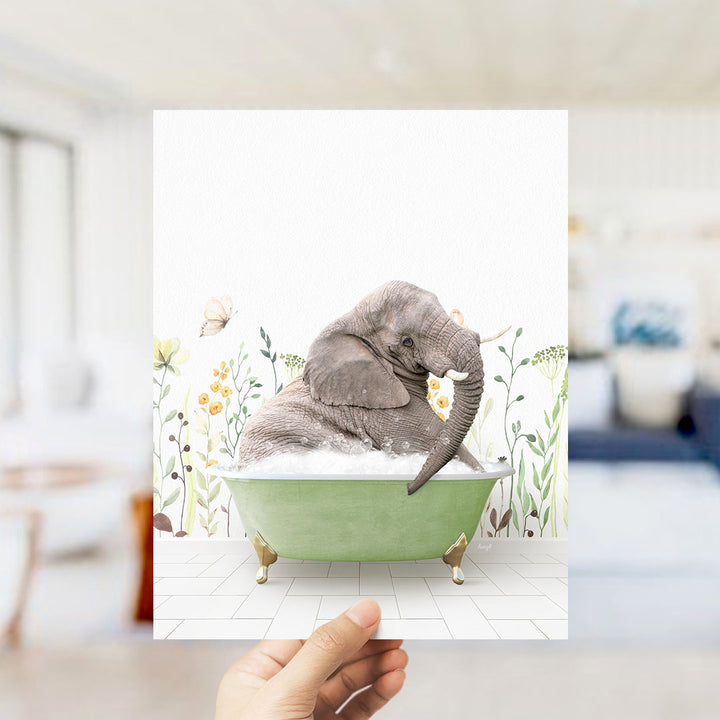 A hand holds up a framed image depicting a gray elephant sitting in a green bathtub filled with bubbles, surrounded by flowers and plants.