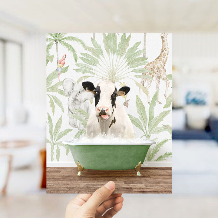 A hand holds up a framed print featuring a cow sitting in a bathtub surrounded by tropical foliage, including a giraffe and palm trees.