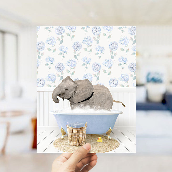 A hand holds up a framed image depicting a gray elephant sitting in a blue bathtub filled with bubbles, surrounded by a floral patterned wall.