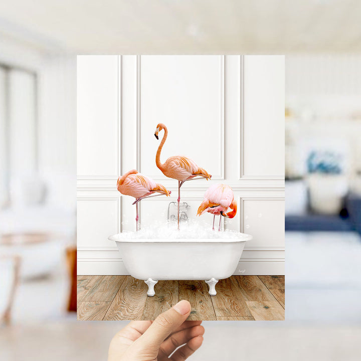 A hand holds up a framed photograph of three flamingos standing in a white bathtub filled with water.