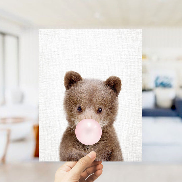 A hand is holding up a square print featuring a cute brown bear blowing a bubble with a pink bubble gum bubble.