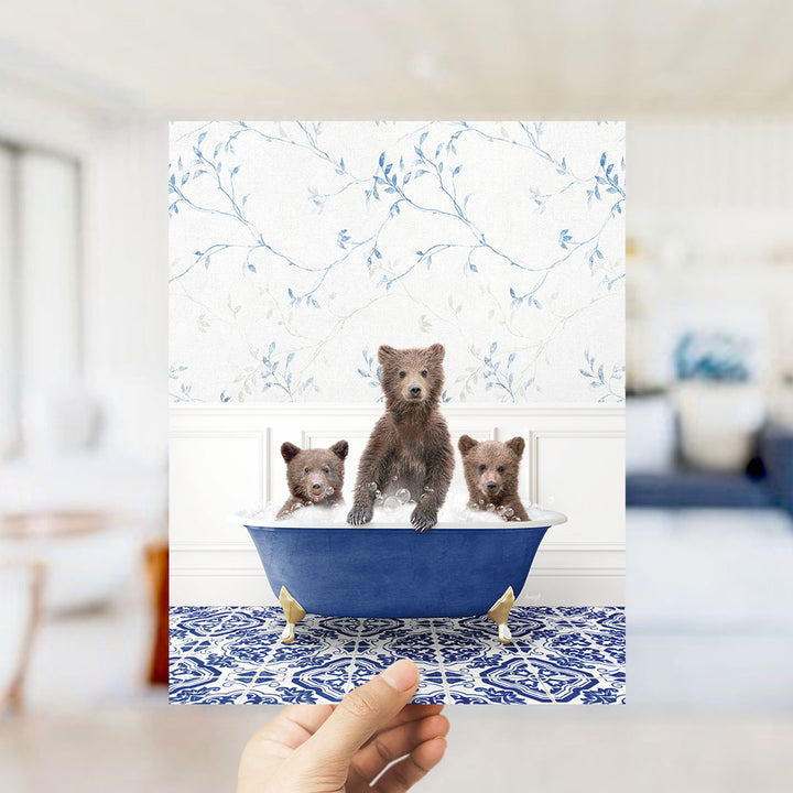 A hand holds up a framed image of three adorable brown bears sitting in a blue bathtub, surrounded by a floral patterned wall.