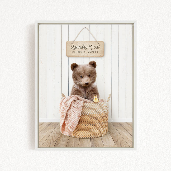 A brown teddy bear sits in a woven basket filled with blankets, with a wooden sign above that reads "Laundry Goal Fluffy Blankets".
