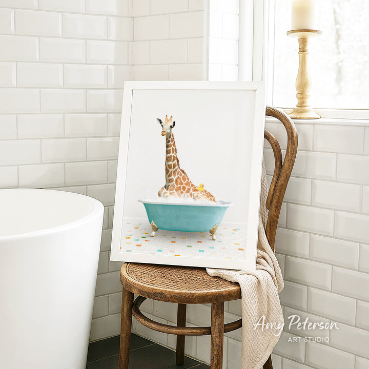 A framed image of a giraffe sitting in a blue bathtub, surrounded by colorful tiles, is displayed on a wooden chair in a bathroom setting.