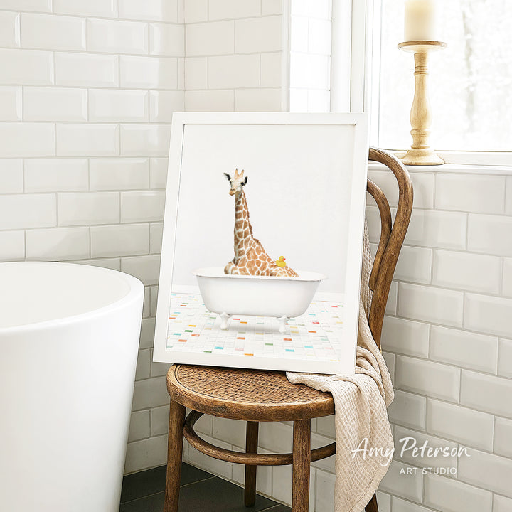 A framed image of a giraffe sitting in a bathtub is displayed on a wooden chair in a bathroom setting.