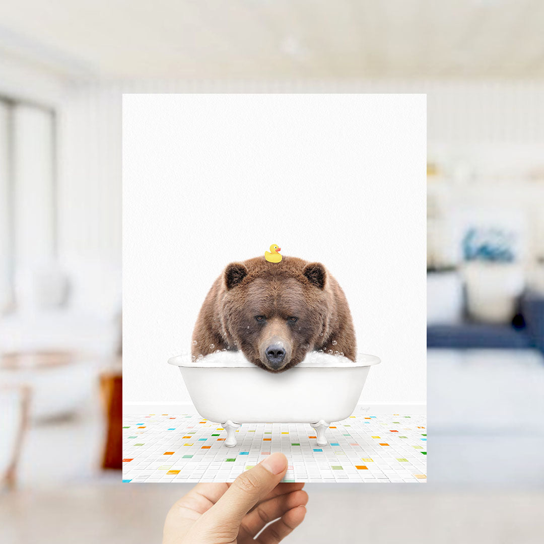 A hand holds up a framed image depicting a brown bear sitting in a white bathtub, with a rubber duck floating on its head.