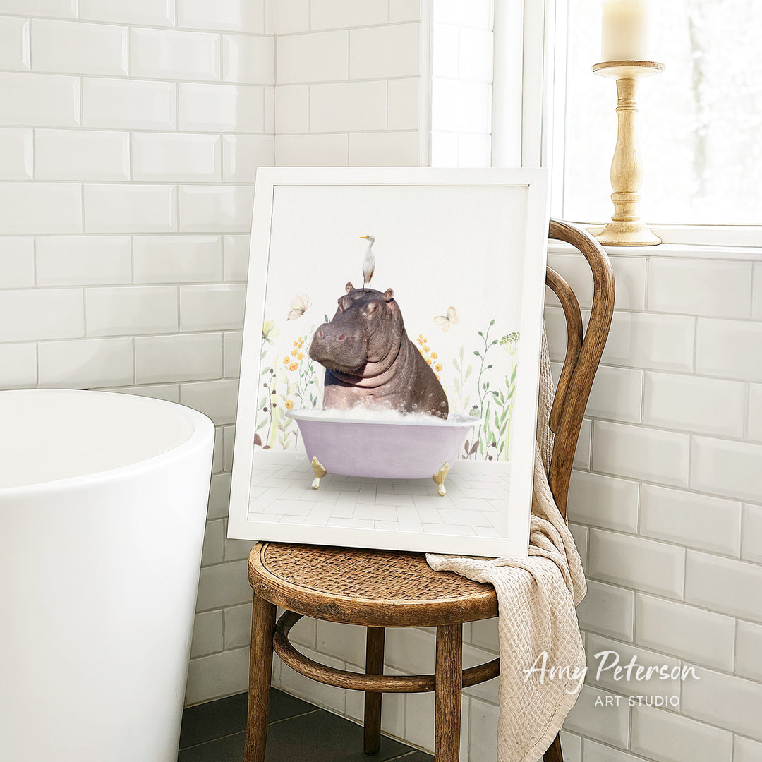 A framed illustration of a hippopotamus sitting in a bathtub, surrounded by flowers and butterflies, is displayed on a wooden chair in a bathroom setting.