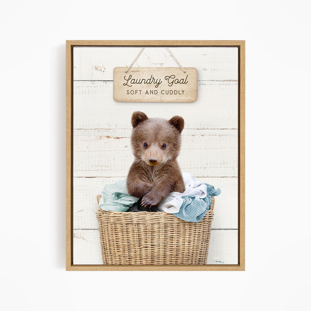 A cute brown bear sitting in a wicker laundry basket, with a wooden sign above that says "Laundry Goal Soft and Cuddly".