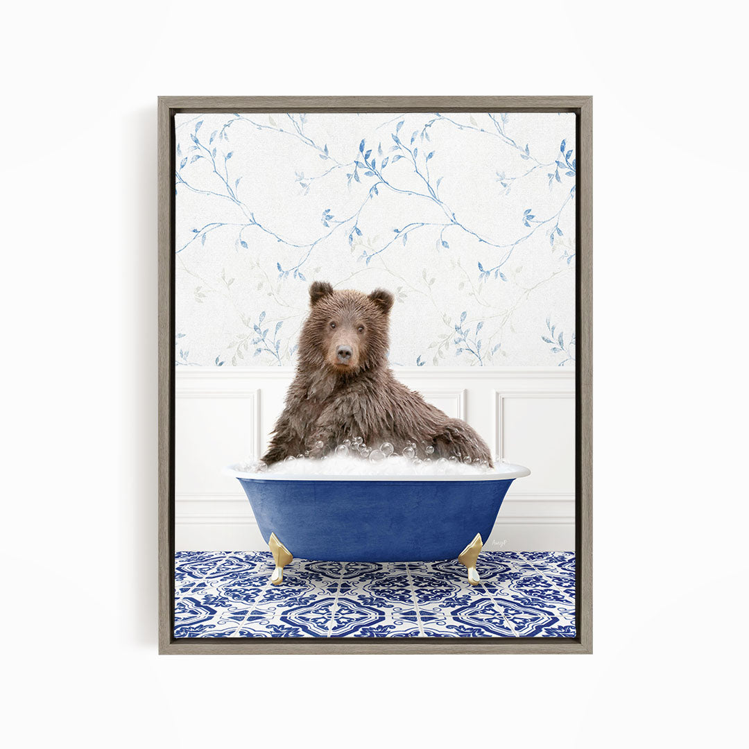 A brown bear sitting in a blue bathtub filled with bubbles, with a blue and white floral patterned wall behind it.