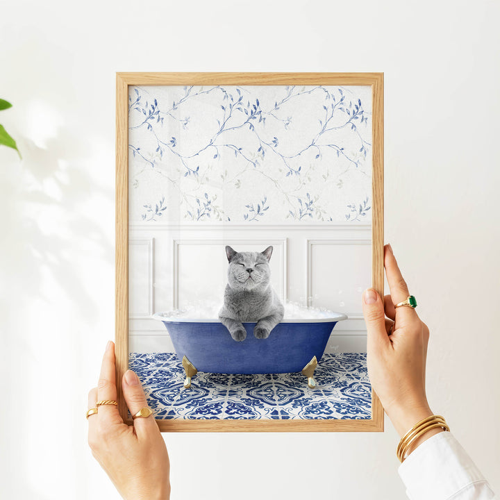 A person's hands are holding a framed picture of a gray cat sitting in a blue bathtub filled with bubbles, with a white floral pattern on the wall behind the cat.