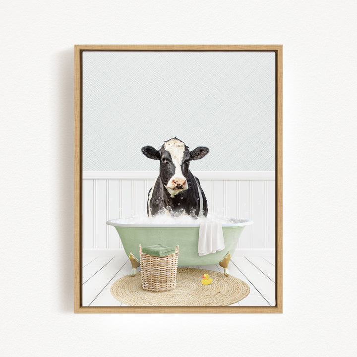 A black and white cow is sitting in a green bathtub filled with water, surrounded by a basket of rubber ducks and a towel.