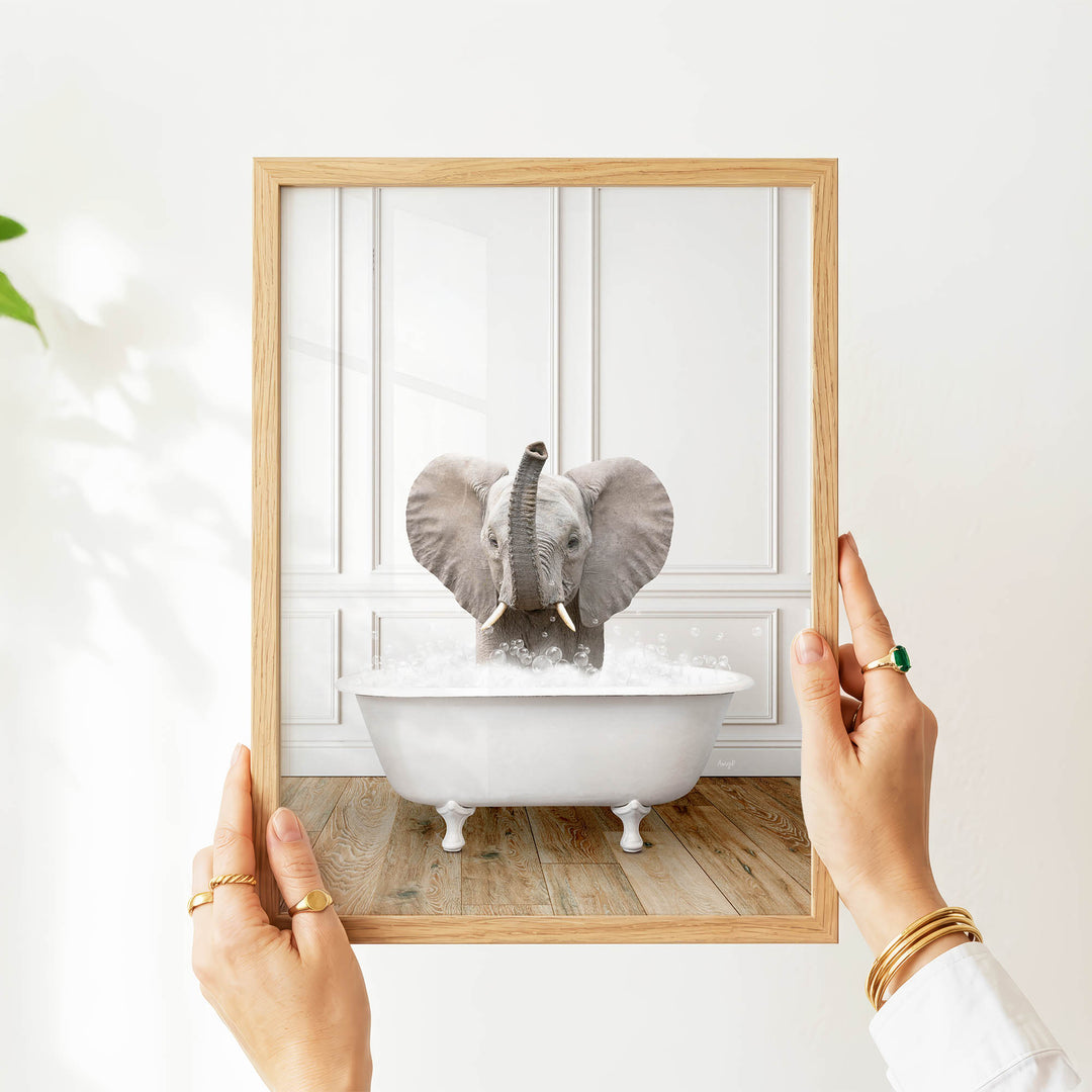 A person's hands are holding a picture frame with a photograph of a baby elephant sitting in a bathtub.