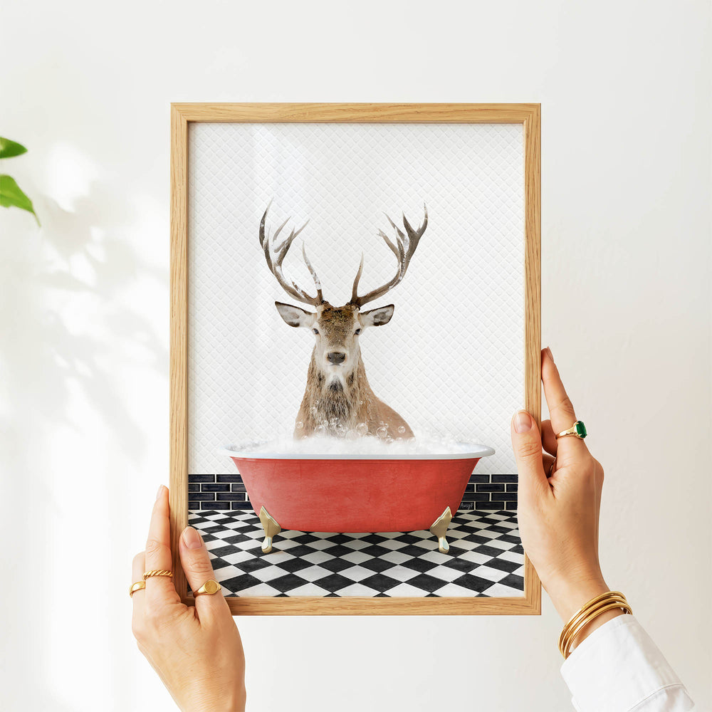 A person's hands are holding a framed picture of a deer in a bathtub, with a black and white checkered floor and a plant in the background.