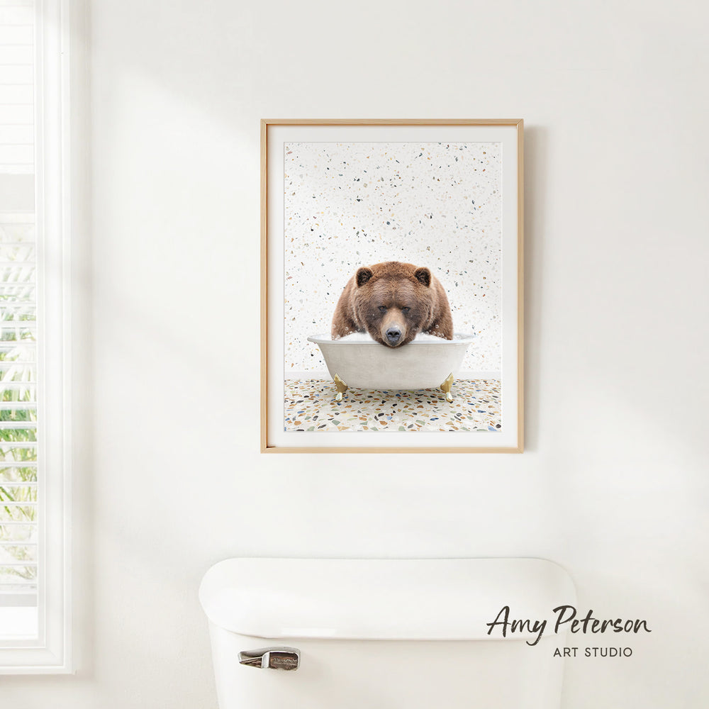 A framed image of a brown bear sitting in a bathtub, surrounded by a speckled background, is hanging on a white wall.