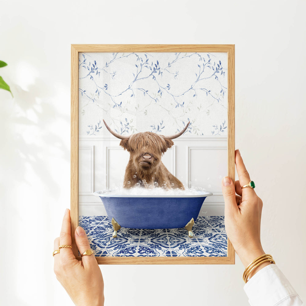 A person's hands are holding a framed picture of a brown cow with long horns sitting in a blue bathtub filled with water.