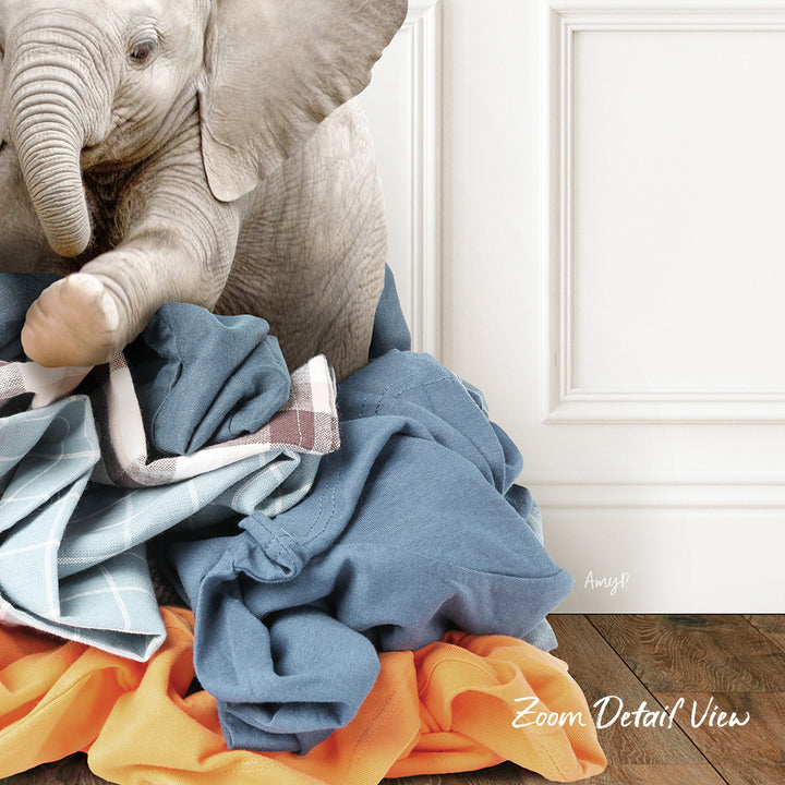 A baby elephant sitting on a pile of clothes, with a white door and wooden floor in the background.