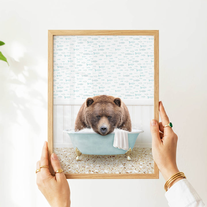 A person's hands are holding a framed image of a brown bear sitting in a bathtub filled with water, surrounded by fish-themed wallpaper.