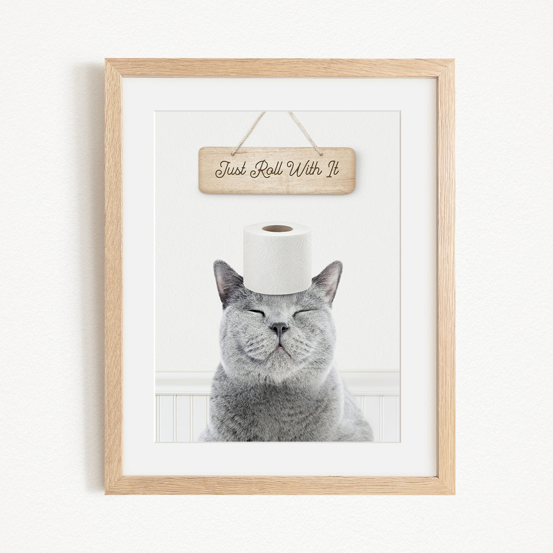 A framed image of a gray cat with its eyes closed, wearing a roll of toilet paper on its head, and a wooden sign above it that says "Just Roll With It".