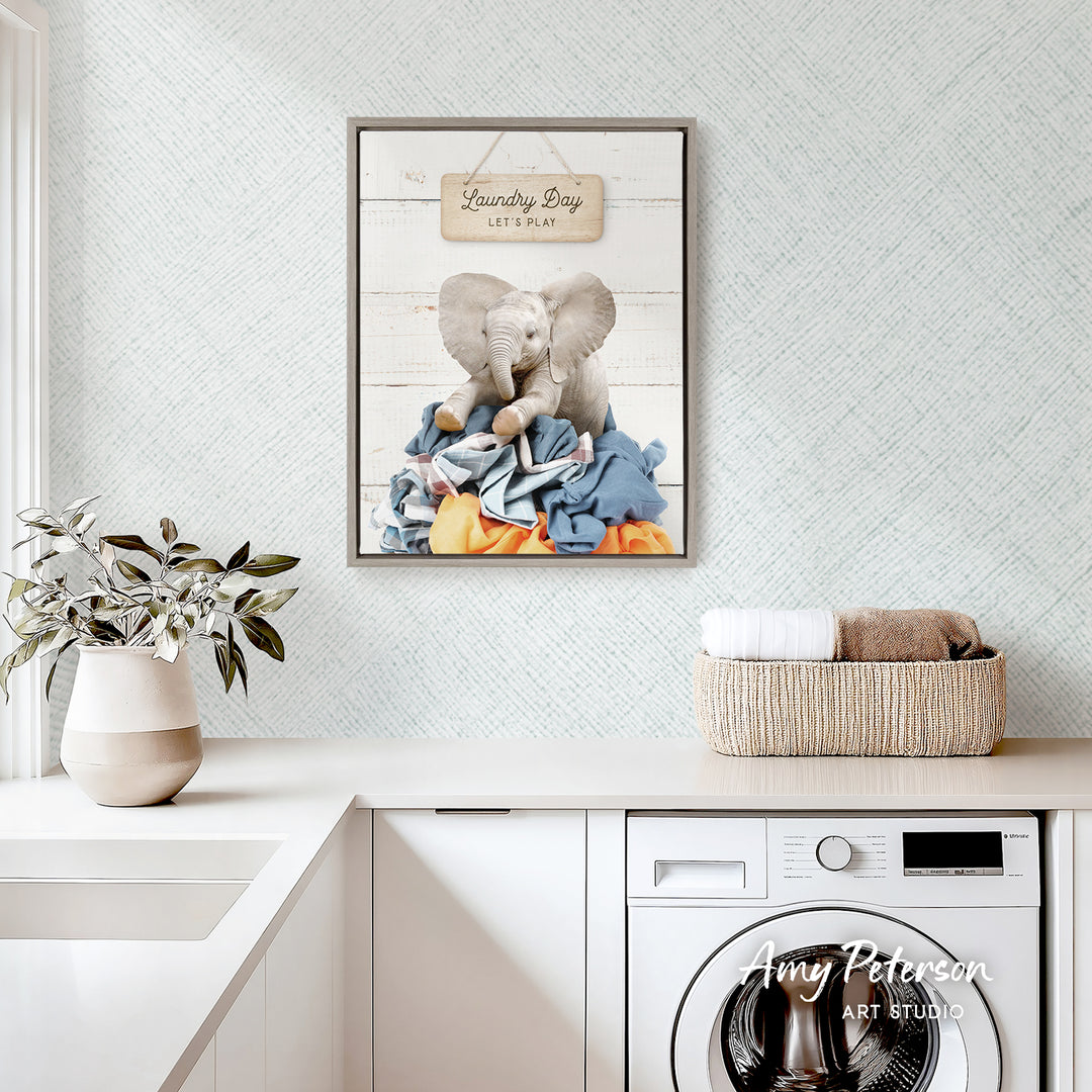 a framed print of an elephant surrounded by laundry, hanging on a wall above a white countertop with a basket of folded clothes and a potted plant.