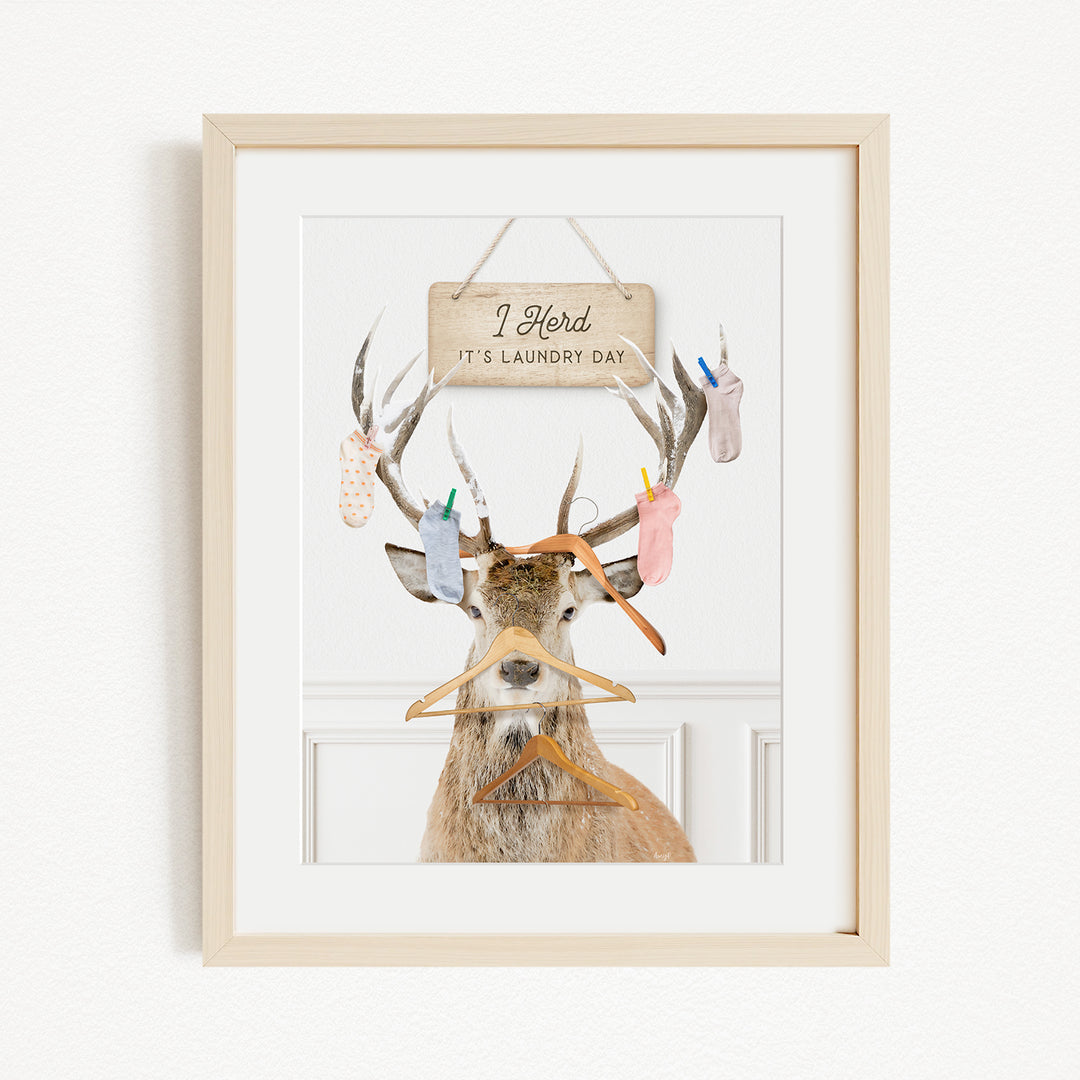 A framed image depicting a deer with antlers holding various laundry items, including socks and clothes hangers, with a sign above that says "I herd its laundry day".