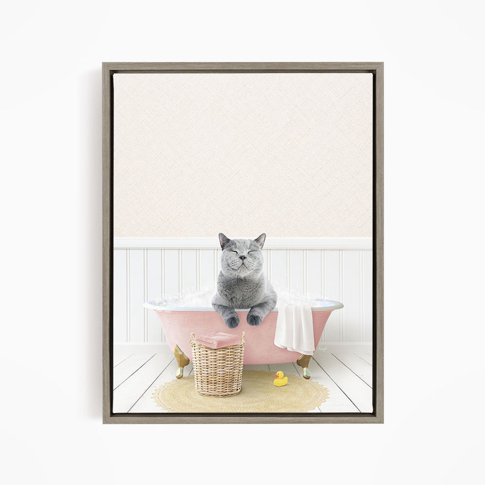 A gray cat sitting in a pink bathtub filled with bubbles, surrounded by a yellow rubber duck and a basket.