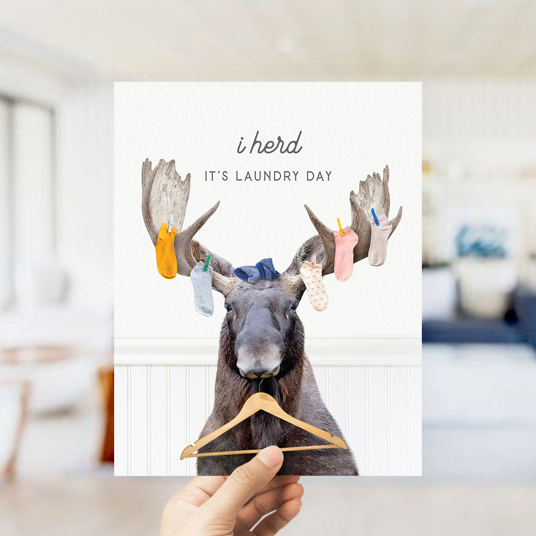 A hand holding a card with a moose wearing clothes and holding a clothesline with clothes hanging on it, along with the text "It's laundry day".