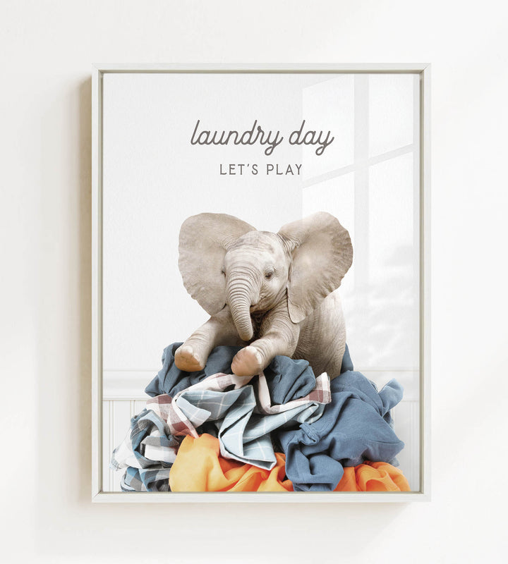 A baby elephant sitting on a pile of laundry, with the text "laundry day let's play" written above it.