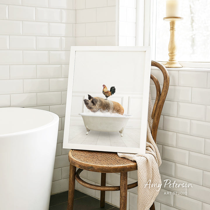 A framed image of a cat sitting in a bathtub, with a rooster perched on the edge of the tub, is displayed on a wooden chair in a bathroom setting.